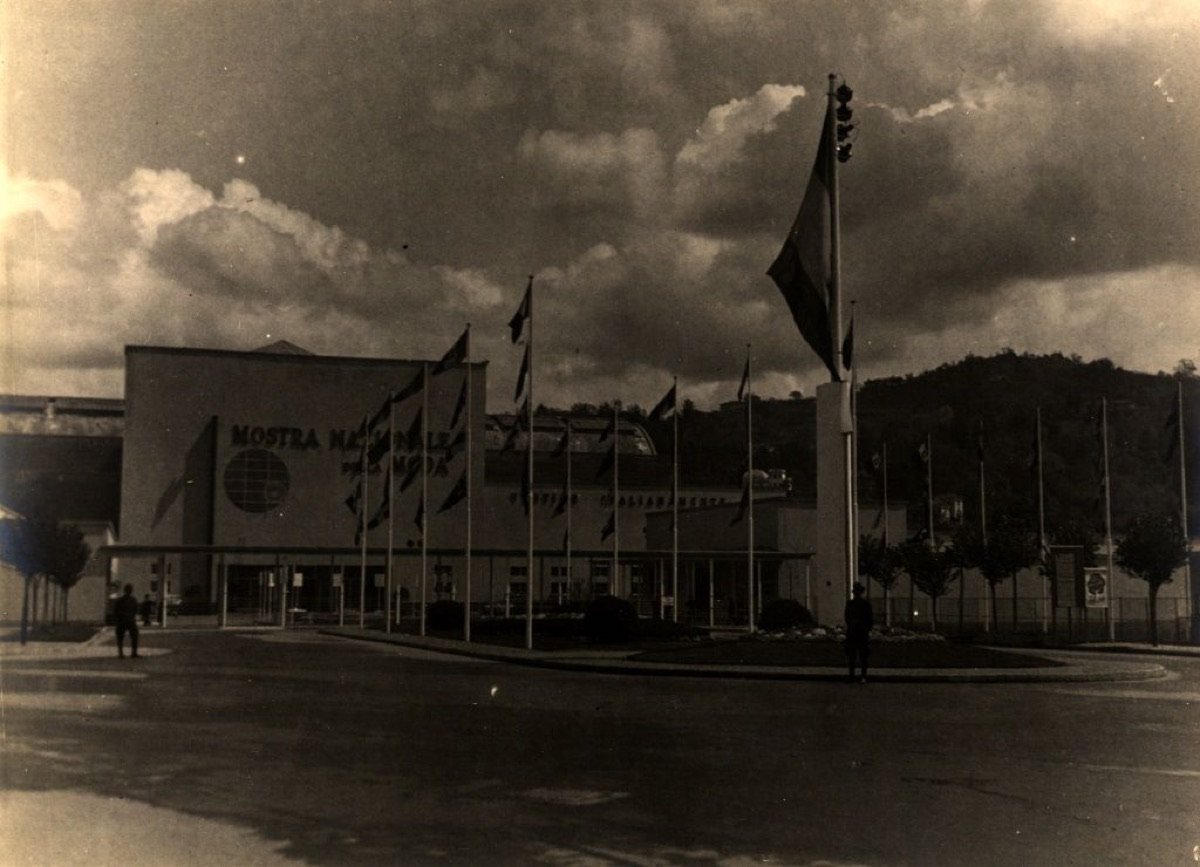 Il Palazzo della Mostra Nazionale della Moda a Torino, edificio modernista con l'insegna «Mostra Nazionale della Moda» e file di bandiere, in una fotografia d'epoca